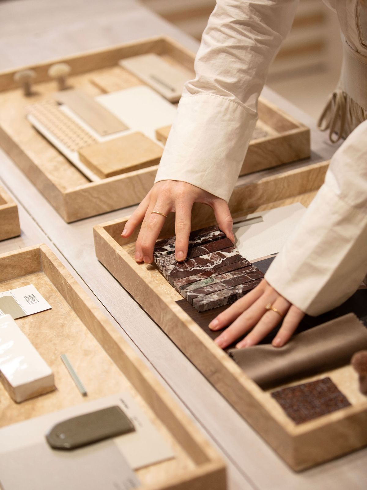A GRID mood board: hands arranging red veined marble strips, dark timber and taupe fabric in travertine trays of stone and finish samples